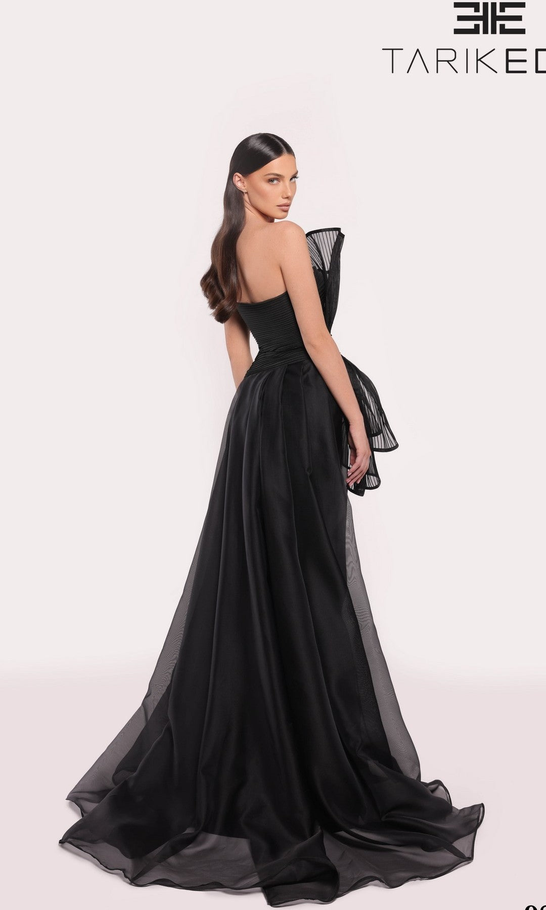 A woman models the Tarik Ediz 98708 Long Formal Dress, an elegant asymmetrical evening gown featuring a large bow detail on the back, styled with sleek hair and neutral makeup against a light background.